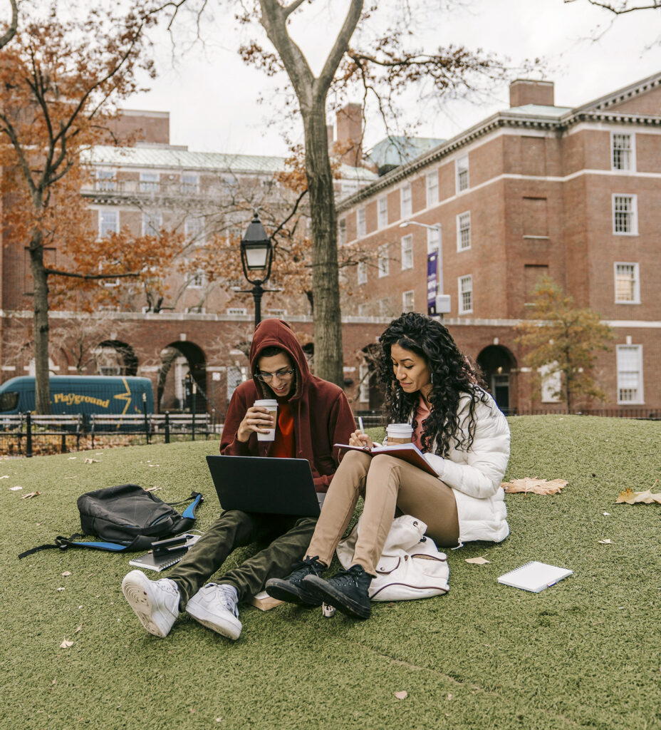 Photo of a female and male college student studying together on a green hill in front of a university, highlighting a relaxed campus atmosphere ideal for focused schoolwork.